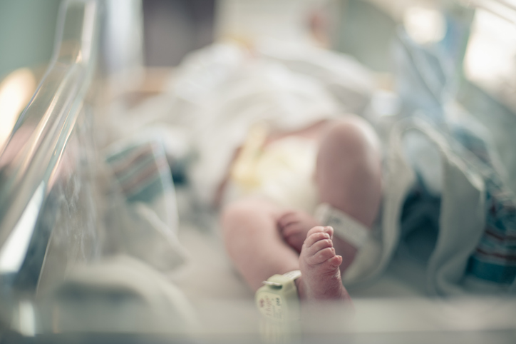 Newborn baby lies on its back surrounded by blankets in hospital bassinet with only its feet and ankle tracking bracelet in focus, photographed with shallow depth of field allowing for ad space.