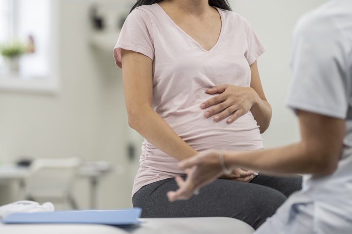 A young pregnant woman, of Asian decent, sits up on an exam table as she talks with her doctor during a prenatal check-up. She is dressed casually and holding her belly as the two talk.
