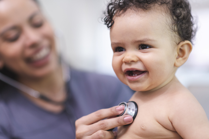 Photo shows a smiling baby seated on a medical exam table.