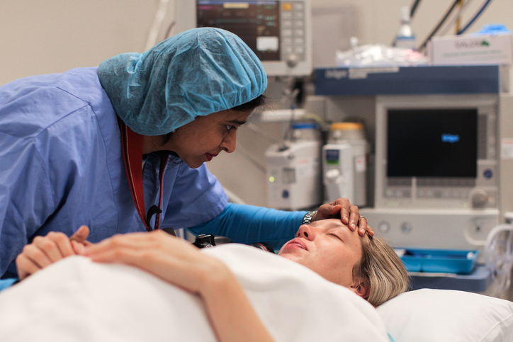 Photo shows a doula supporting her pregnant client as she lays in the operating room.