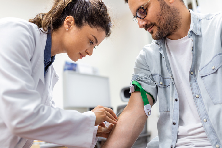 Photo shows a phlebotomist collecting blood sample from a male patient in a lab/Getty Images