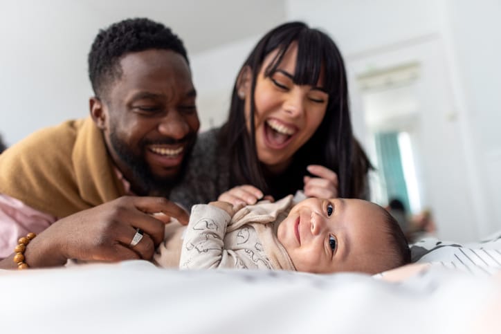 Photo shows a mother and father laughing and overlooking their newborn baby lying on a bed/Getty Images